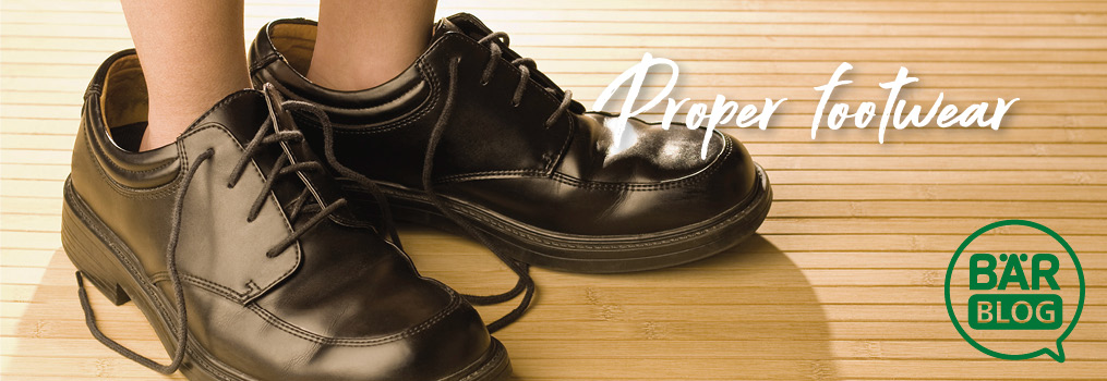 Person wearing oversized black leather shoes on wooden floor with 'Proper Footwear' text