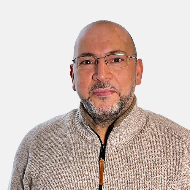 Ein Mann trägt eine Brille und einen beigen Pullover mit Reißverschluss, vor einem neutralen Hintergrund