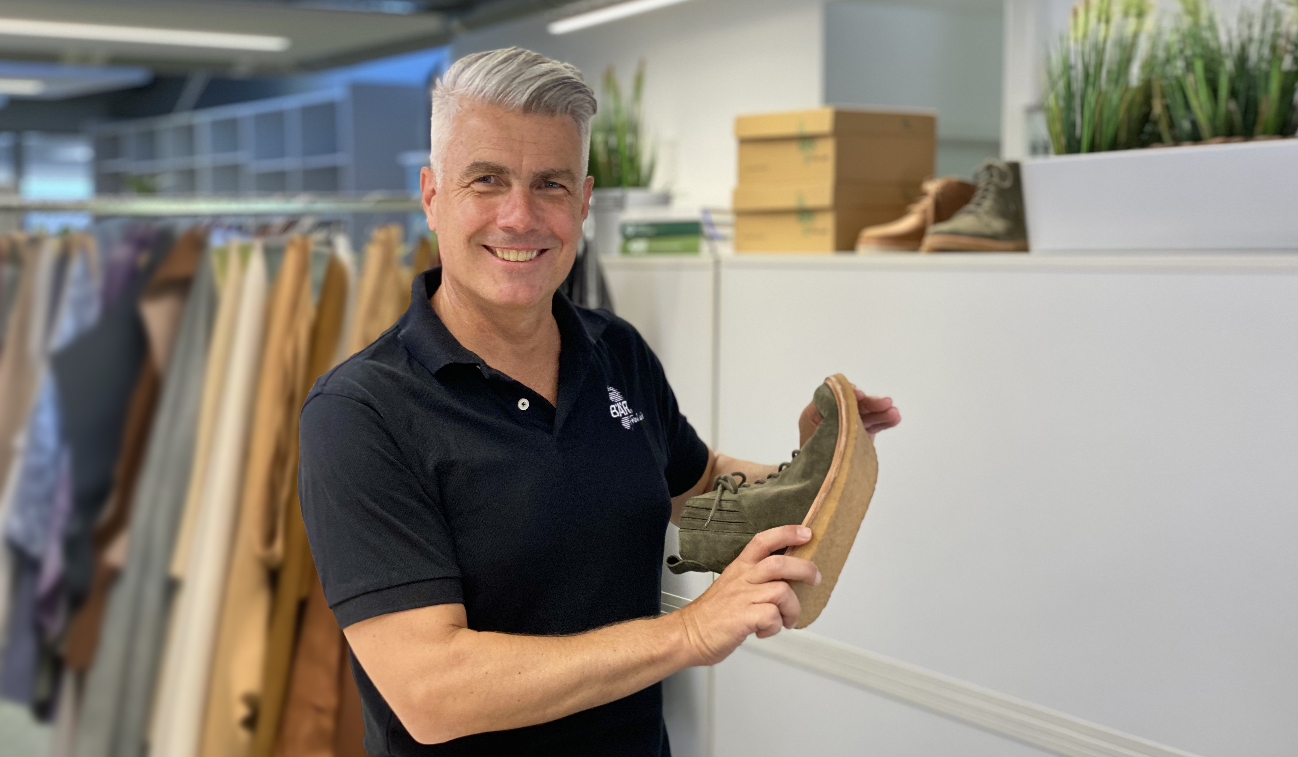 Mann in schwarzem Polo zeigt grünen Schuh in Büro Mann mit grauen Haaren in schwarzem Polohemd hält grünen Schuh im Büro mit Kleiderständern im Hintergrund
