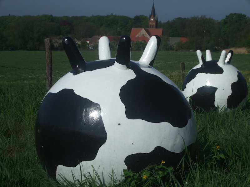 Round, black and white cow-patterned sculptures in a grassy field with a village and church in the background