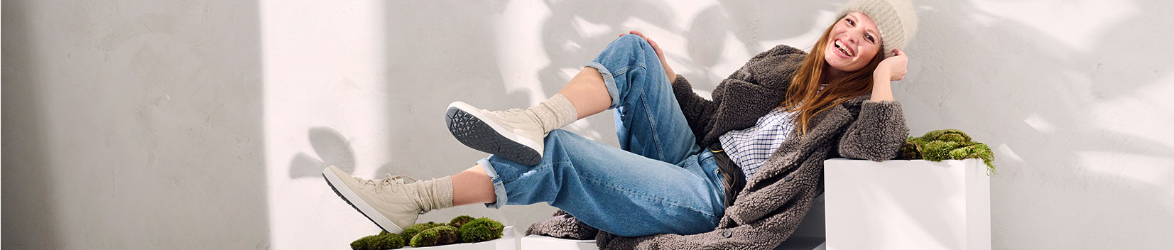 Cozy Winter Outfit with Knit Beanie and Casual Sneakers Woman in a warm coat and knit beanie relaxing in casual sneakers and jeans, surrounded by white decor and greenery.