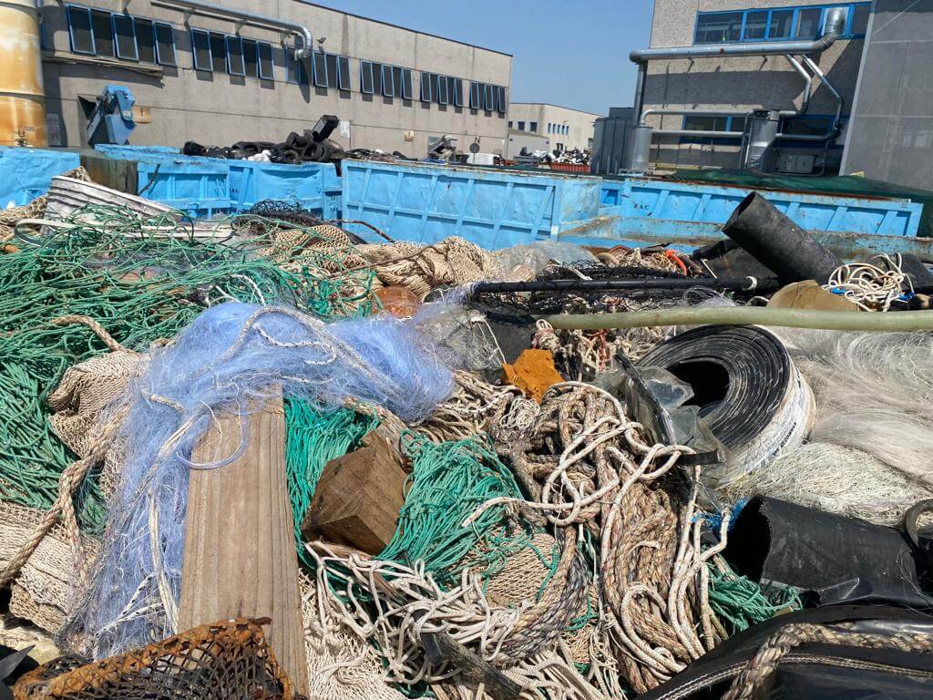 A heap of tangled fishing nets, ropes, and other materials at a recycling center with industrial buildings in the background.