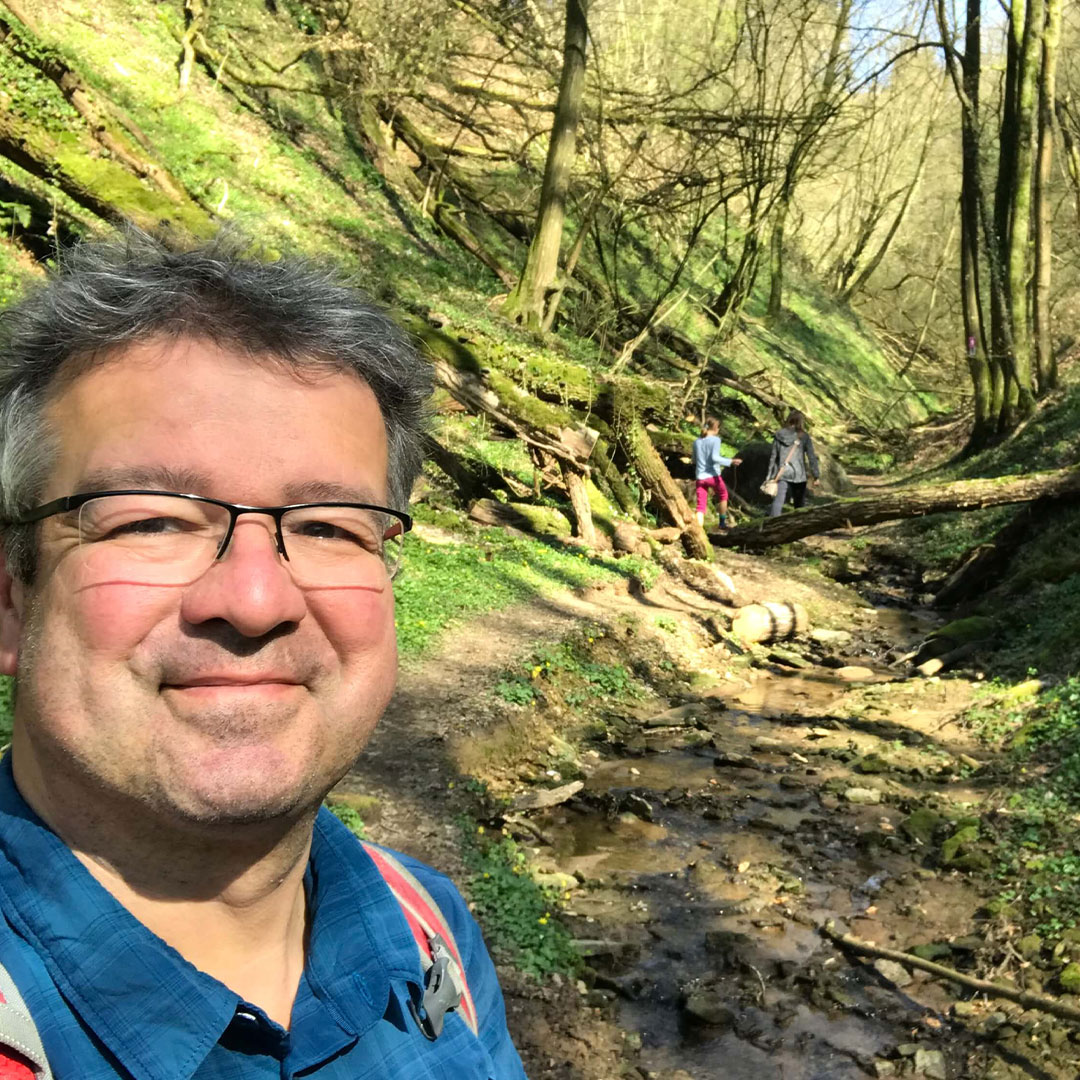 Wanderer auf einem Waldweg mit Bach Ein Mann mit Brille und Rucksack steht auf einem schattigen Waldweg neben einem kleinen Bach, weitere Wanderer sind im Hintergrund zu sehen.
