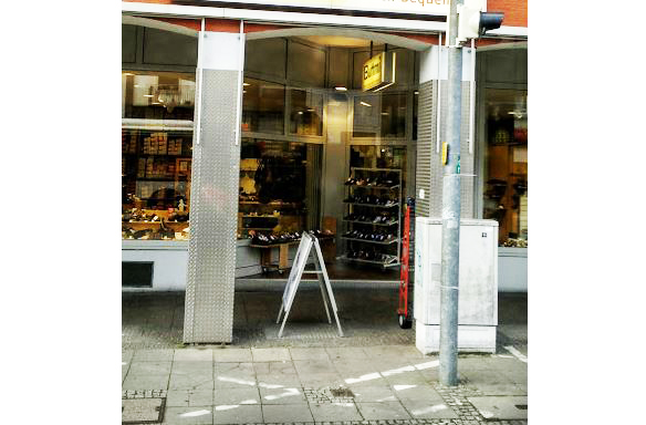 Eine Straße mit Blick auf die Fensterfront eines Schuhgeschäfts, wo im Inneren Schuhe auf Regalen ausgestellt sind.