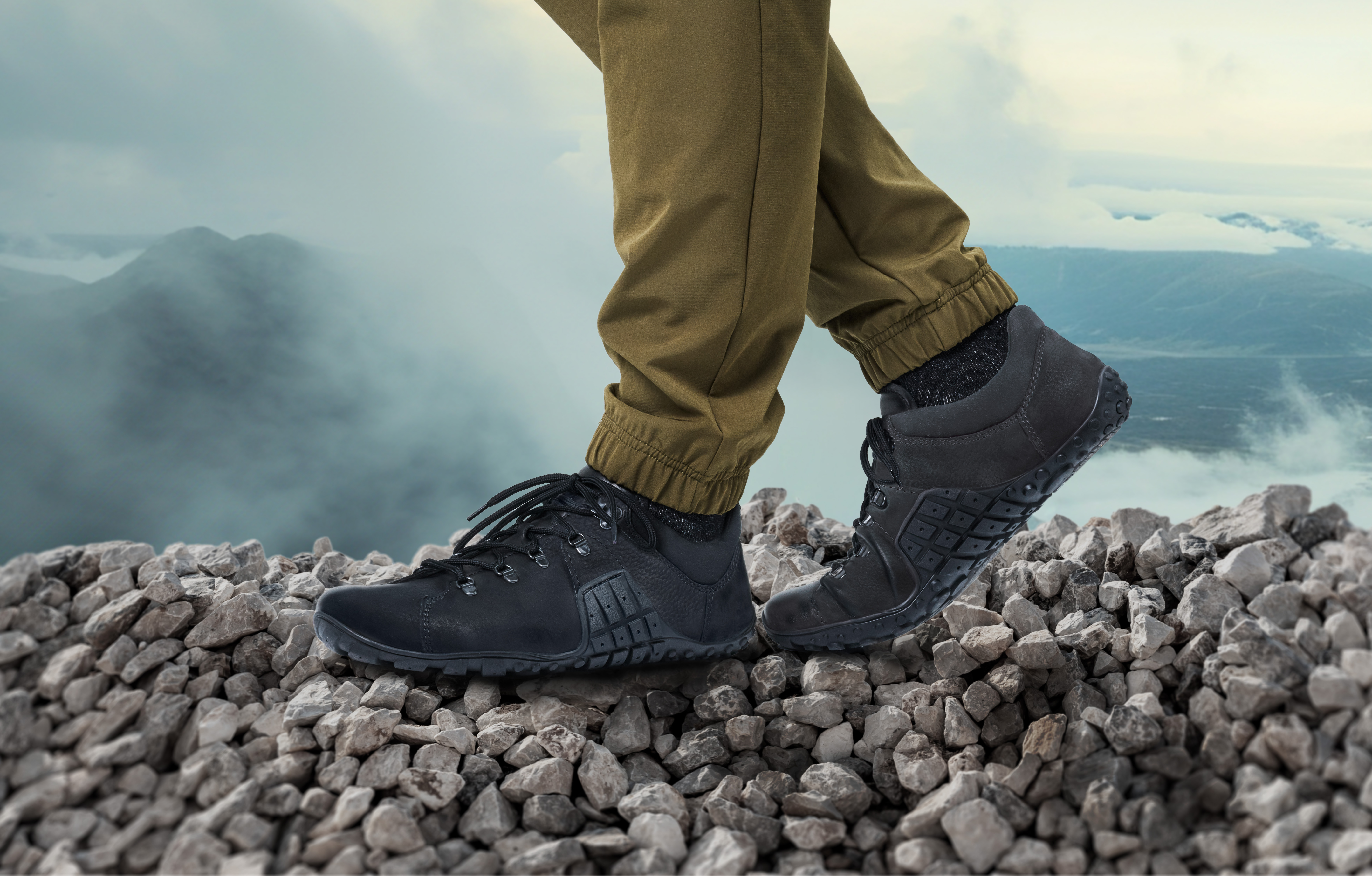 Outdoor-Wanderschuhe auf steinigem Untergrund vor nebligem Bergpanorama Person in Outdoor-Hosen trägt schwarze Wanderschuhe und steht auf einem steinigen Pfad mit Bergen im Hintergrund