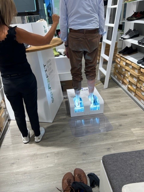 Analyse der Fußstellung mit moderner Technologie im Schuhgeschäft Ein Mann steht auf einem Gerät, das seine Füße scannt, während eine Verkäuferin daneben steht. Umgeben von Regalen mit Schuhkartons.