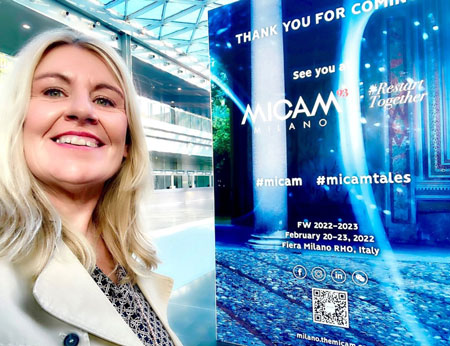 Visitor at MICAM Milano Event A woman smiling next to a MICAM Milano event poster.
