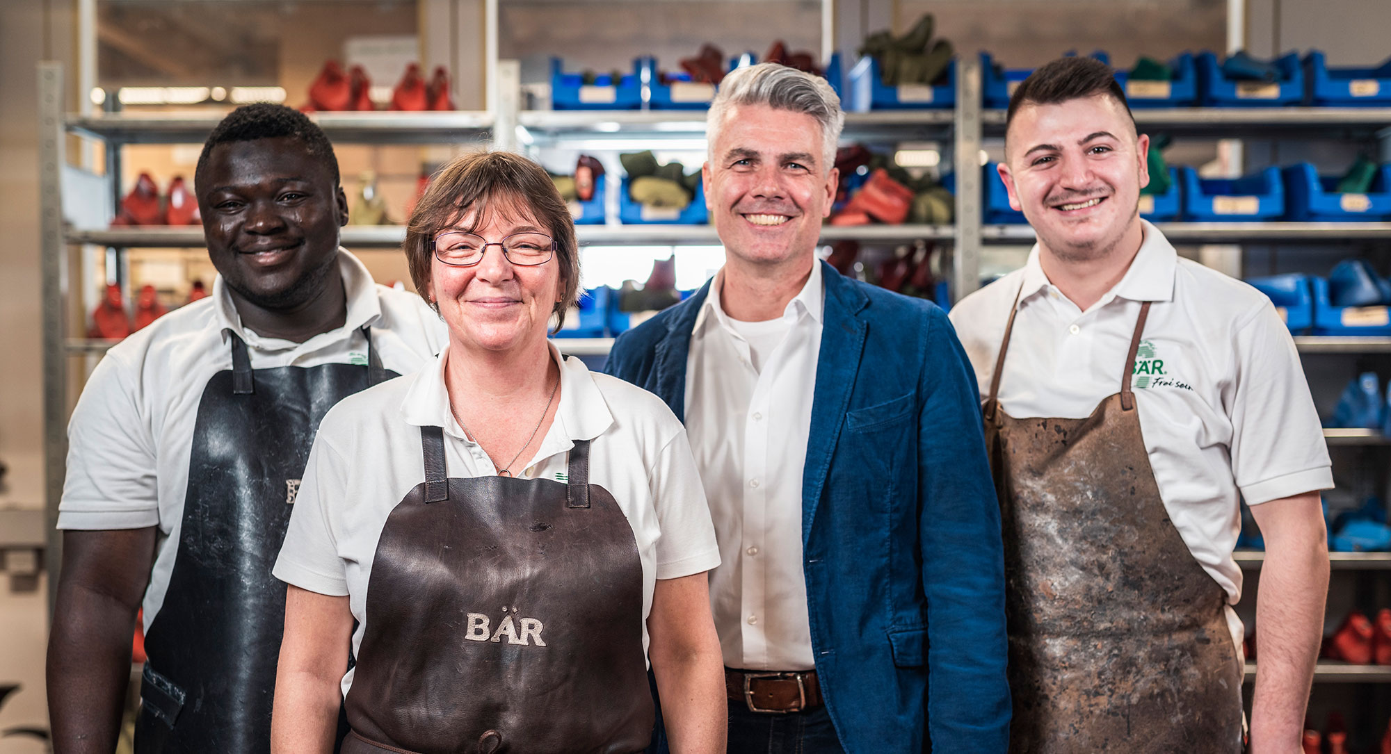 Team der Schuhmanufaktur im Arbeitsbereich Vier Personen in Arbeitskleidung in einer Schuhmanufaktur stehen lächelnd vor Regalen mit Schuhen.
