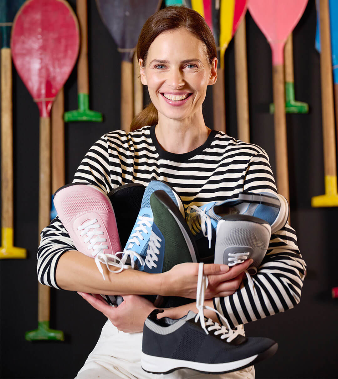 Smiling woman holding colorful sneakers in front of paddle background A woman in a striped shirt smiles while holding several pairs of colorful sneakers with paddles in the background.