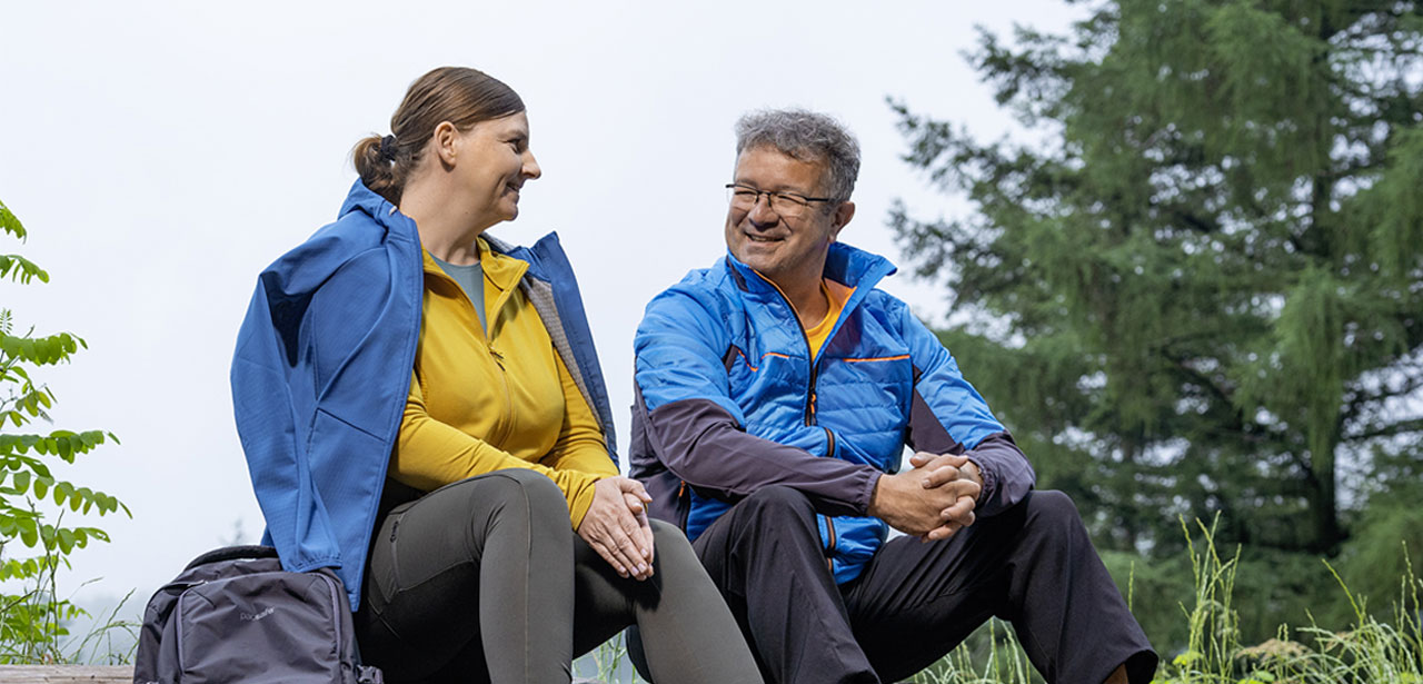 Couple Enjoying a Nature Hike in Outdoor Gear A man and a woman sitting on a log in a forest, wearing blue and yellow outdoor jackets, smiling at each other.