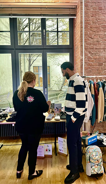 Zwei Personen bei der Auswahl von Schuhen im Modegeschäft Eine Frau und ein Mann betrachten Schuhe in einem Modegeschäft. Im Hintergrund sind ein Fenster und ein Kleiderständer mit Jacken sichtbar.