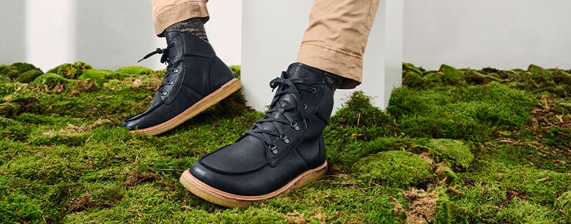 Schwarz gekleidete Lederstiefel auf grünem Moos mit beigen Hosenbeinen
