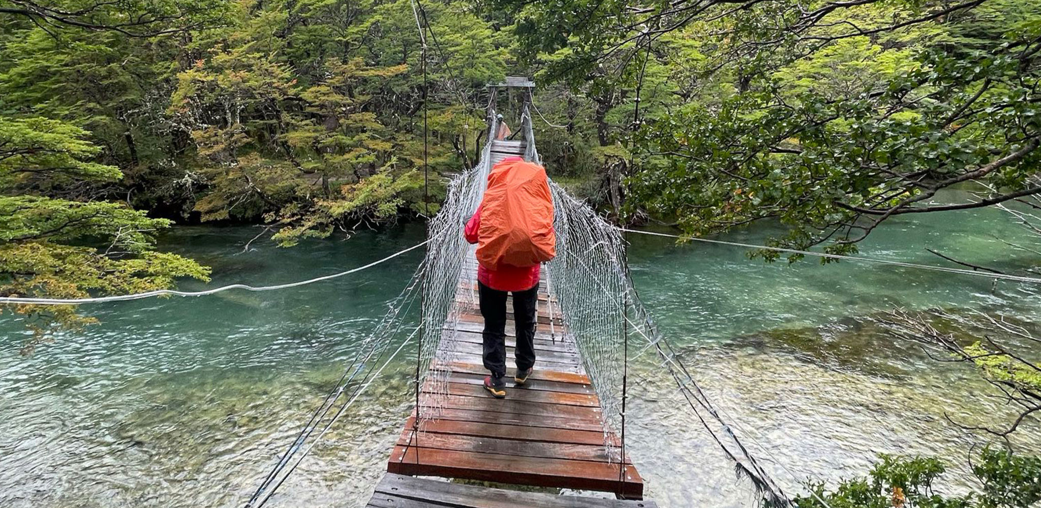 Hiker crossing a wooden suspension bridge over a clear river in a lush forest A person with an orange backpack walks on a wooden suspension bridge across a clear, green-tinted river, surrounded by dense green forest.