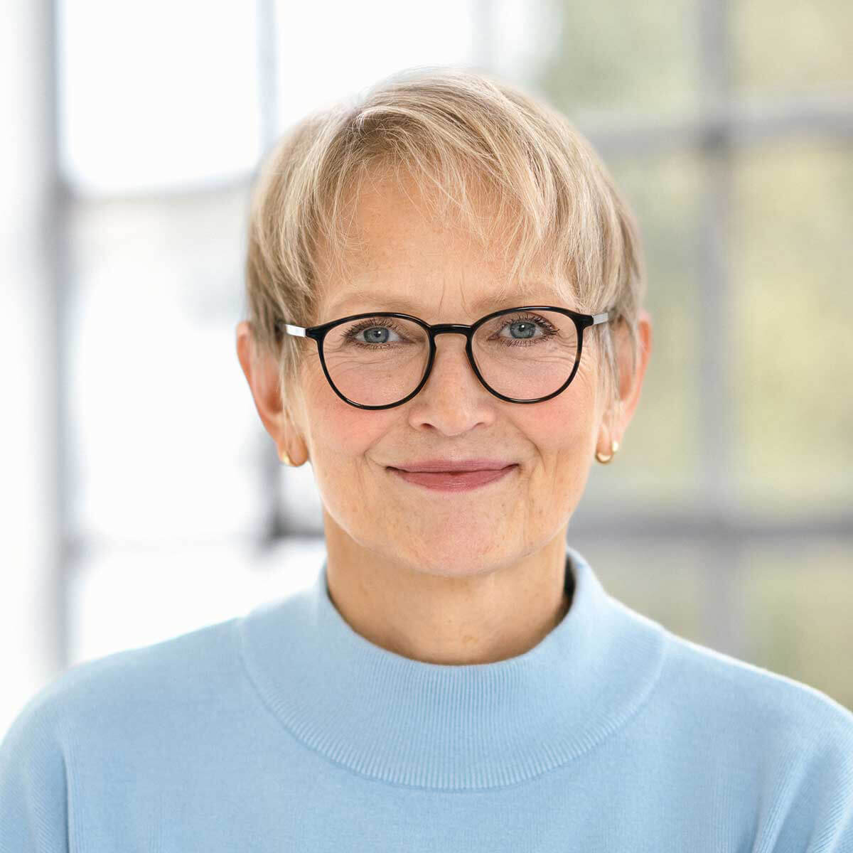 Portrait of a smiling woman with glasses in a light blue sweater A woman with short blonde hair and glasses wearing a light blue sweater, smiling in front of a window