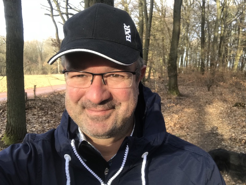 A man wearing a cap and glasses smiling in a sunlit forest
