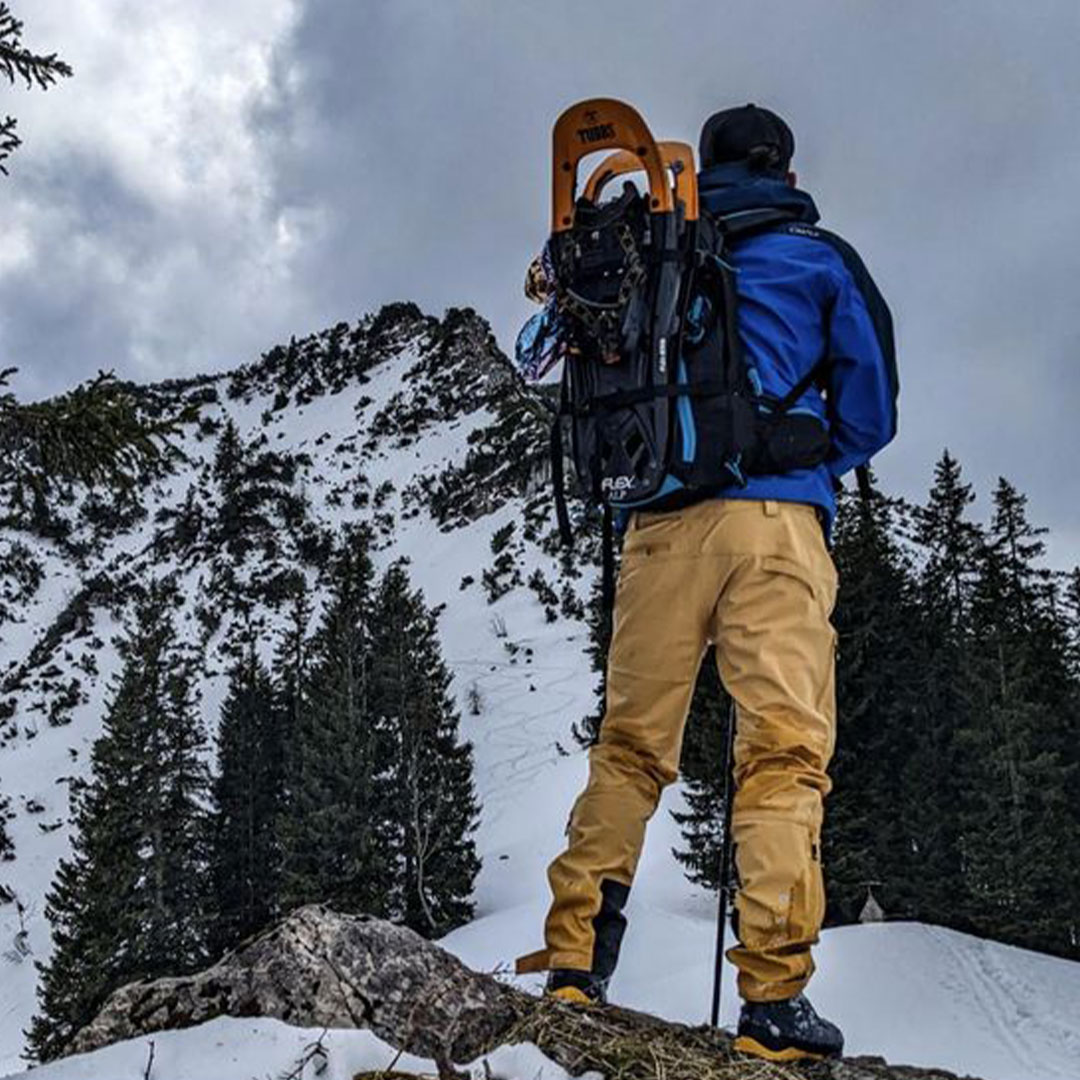Mann in blauer Jacke und gelber Hose mit Schneeschuhen im Rucksack vor verschneiter Bergkulisse